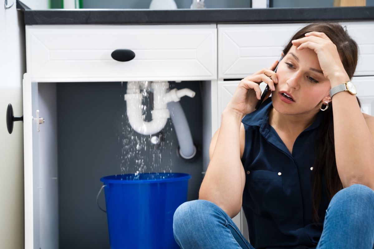 A woman on the phone looks stressed as water leaks from a pipe under the kitchen sink into a bucket, worried that the damage might extend to the bathroom subfloor.