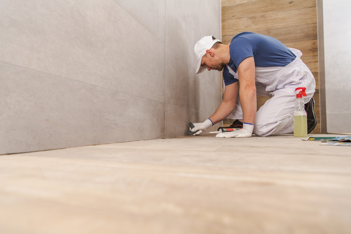 A worker in white overalls and a blue shirt crouches on the bathroom subfloor, carefully applying grout between tiles. A spray bottle and other tools are beside him.