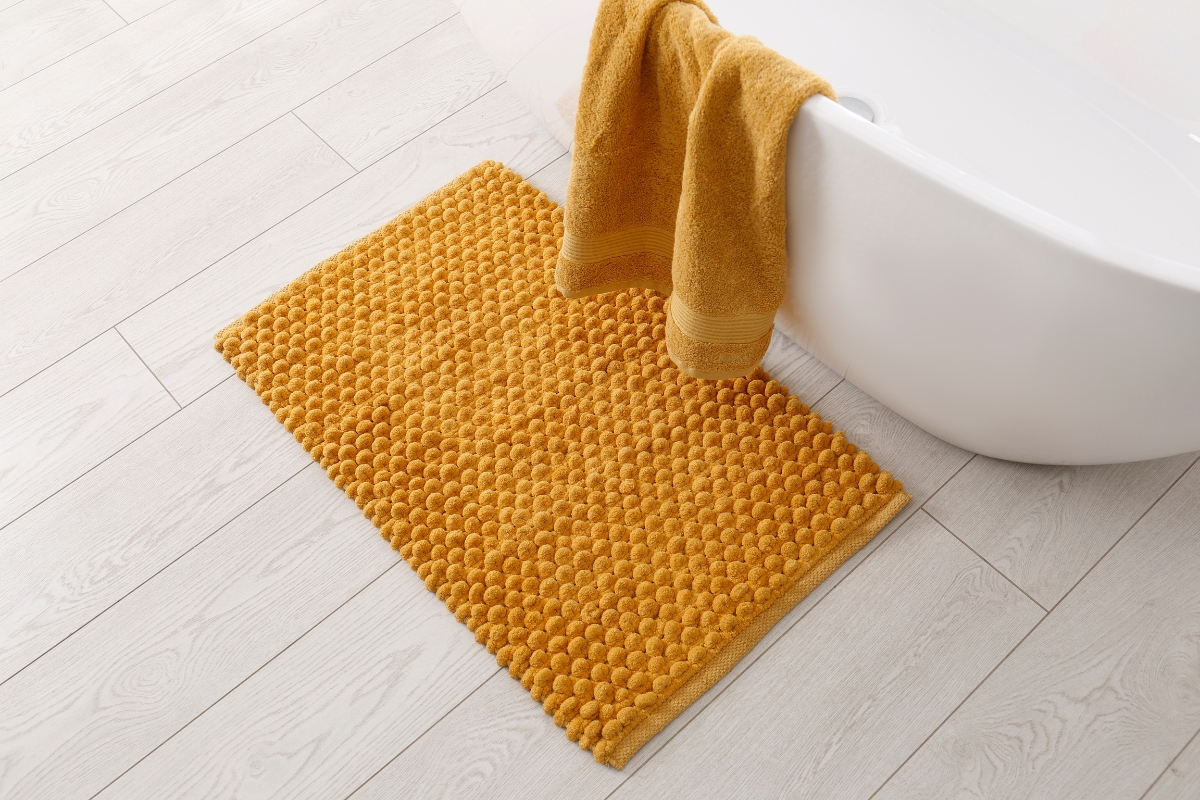 A mustard yellow bath mat with a textured pattern is placed on a light wood bathroom subfloor beside a white bathtub, with a matching yellow towel hanging over the rim.