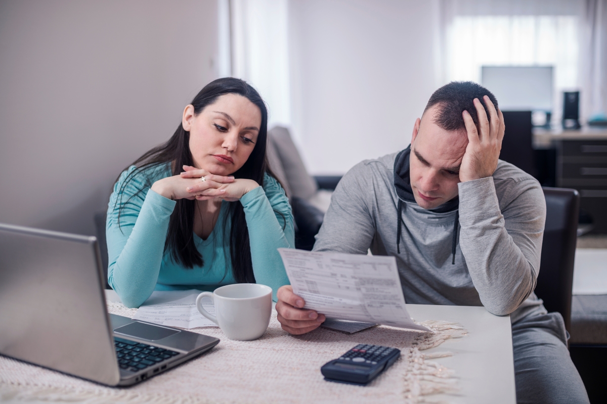 A couple looks concerned while reviewing documents, likely regarding their bathroom subfloor, at a table with a laptop, a coffee mug, and a calculator.