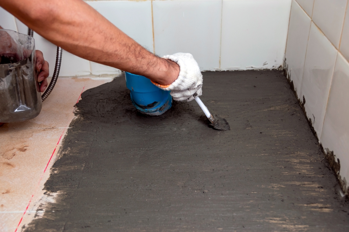 A person wearing a white glove uses a trowel to spread mortar or grout on a tiled bathroom subfloor, with white tiles on the wall in the background.