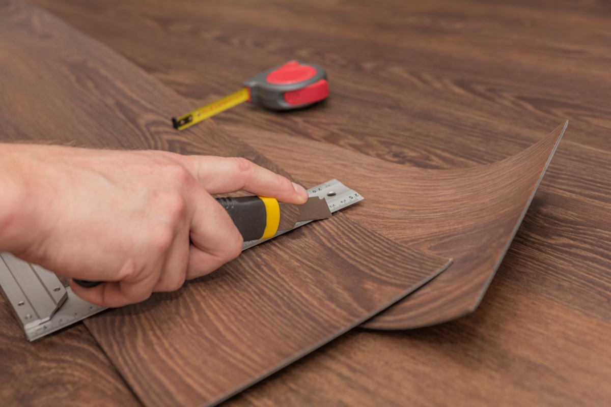 A person cutting glue down vinyl plank flooring using a utility knife and a metal ruler with a tape measure in the background.