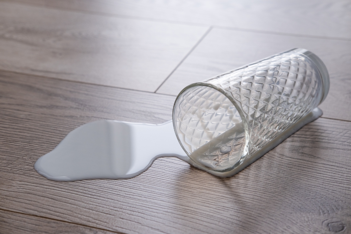 A glass lying on its side on glue down vinyl plank flooring with spilled milk coming out of it.