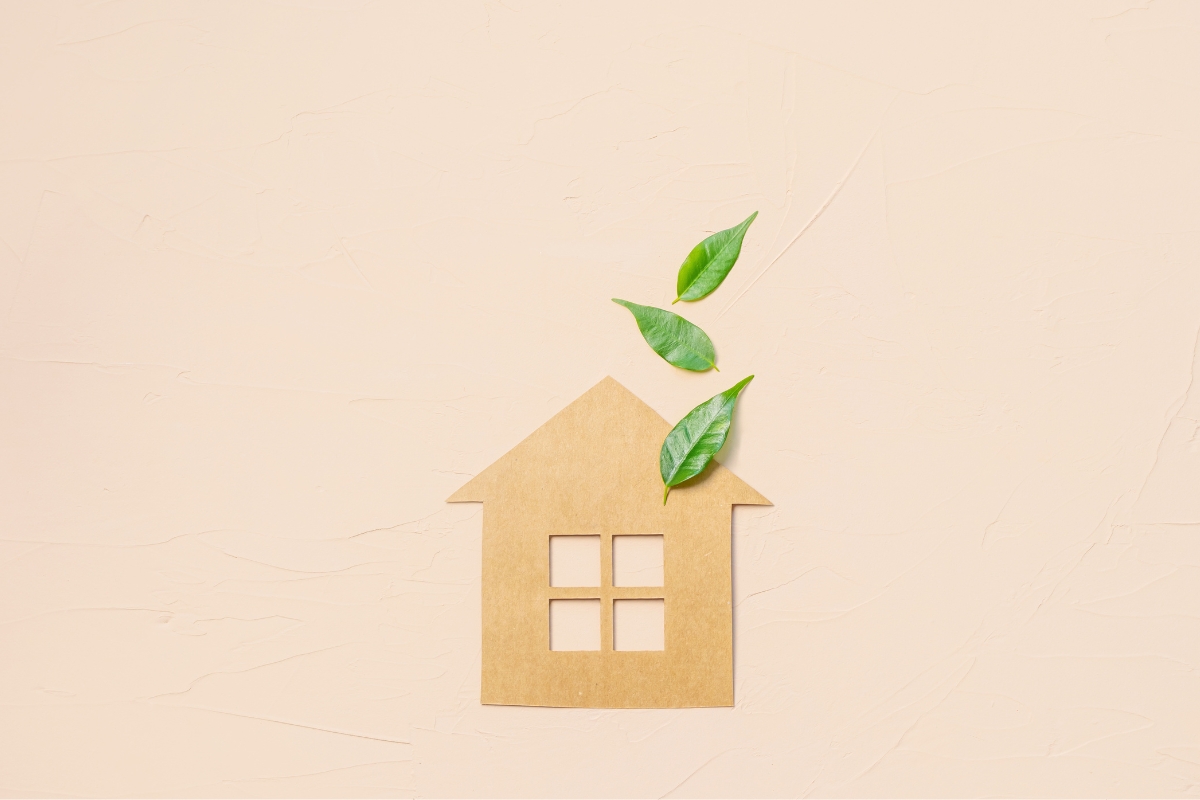 A cardboard cutout of a house with four windows is placed on a flat surface, featuring glue down vinyl plank flooring. Three green leaves are positioned above the roof, suggesting the concept of eco-friendly housing.