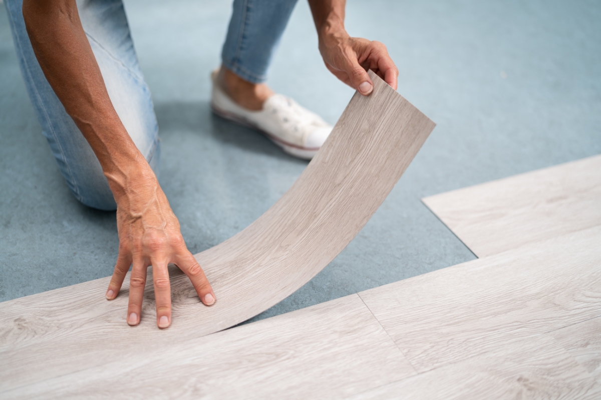 A person is installing glue down vinyl plank flooring, carefully bending a piece into place on a blue underlayment. Wearing jeans and white sneakers, they ensure each plank fits perfectly.