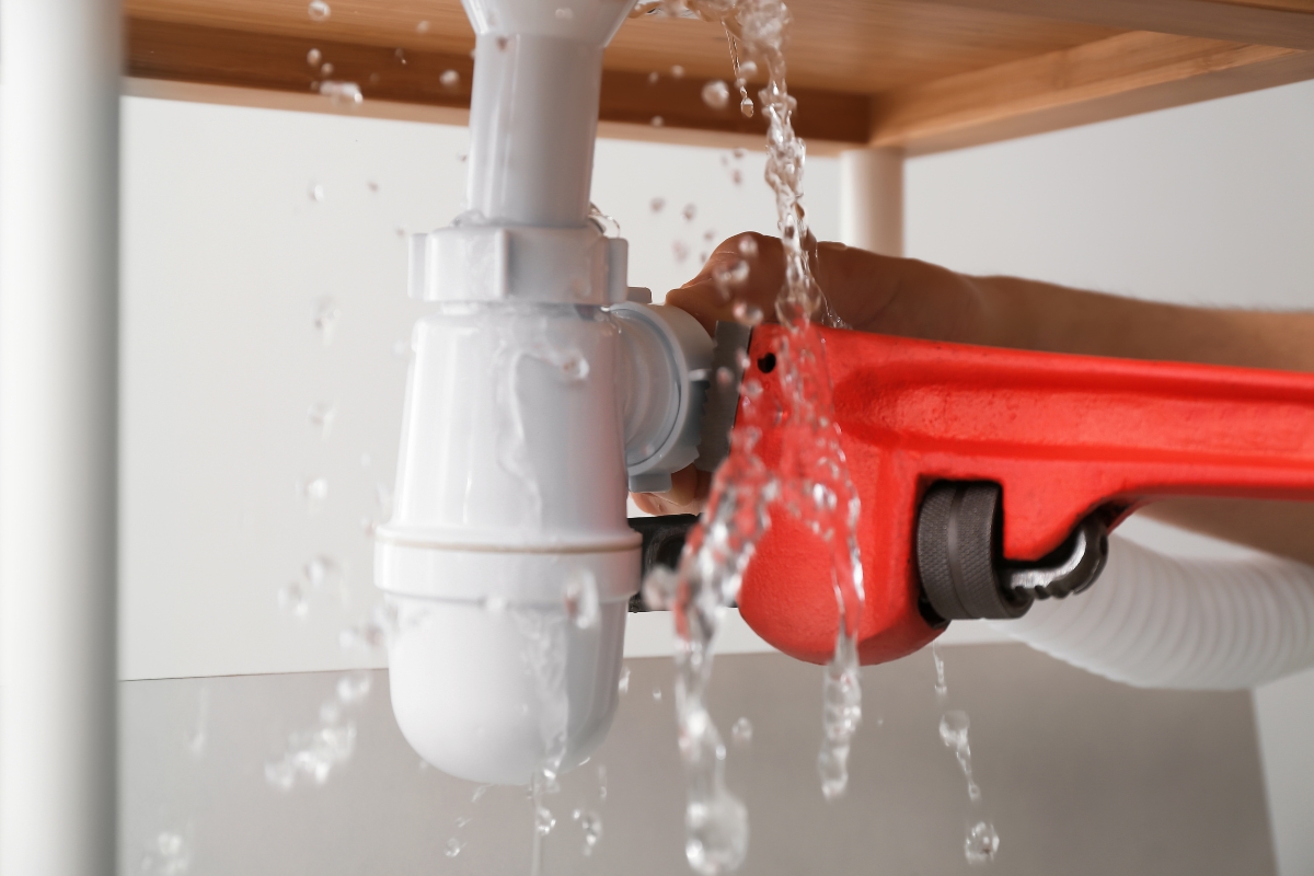 A person uses a red wrench to fix a leaking white pipe under a sink, with water pouring out, diligently preventing water damage.