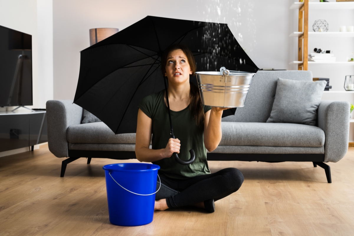 A woman sits on the floor of a living room holding a bucket under a leaking ceiling while also using an umbrella, preventing water damage. A blue bucket is placed beside her on the floor.