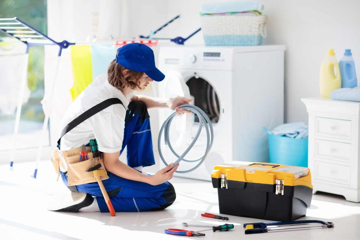 A person in a blue uniform and cap, with a tool belt, kneels in front of a washing machine checking a hose, ensuring everything's secure to prevent water damage. A toolbox and various tools are scattered nearby. Laundry items and detergent bottles are in the background.