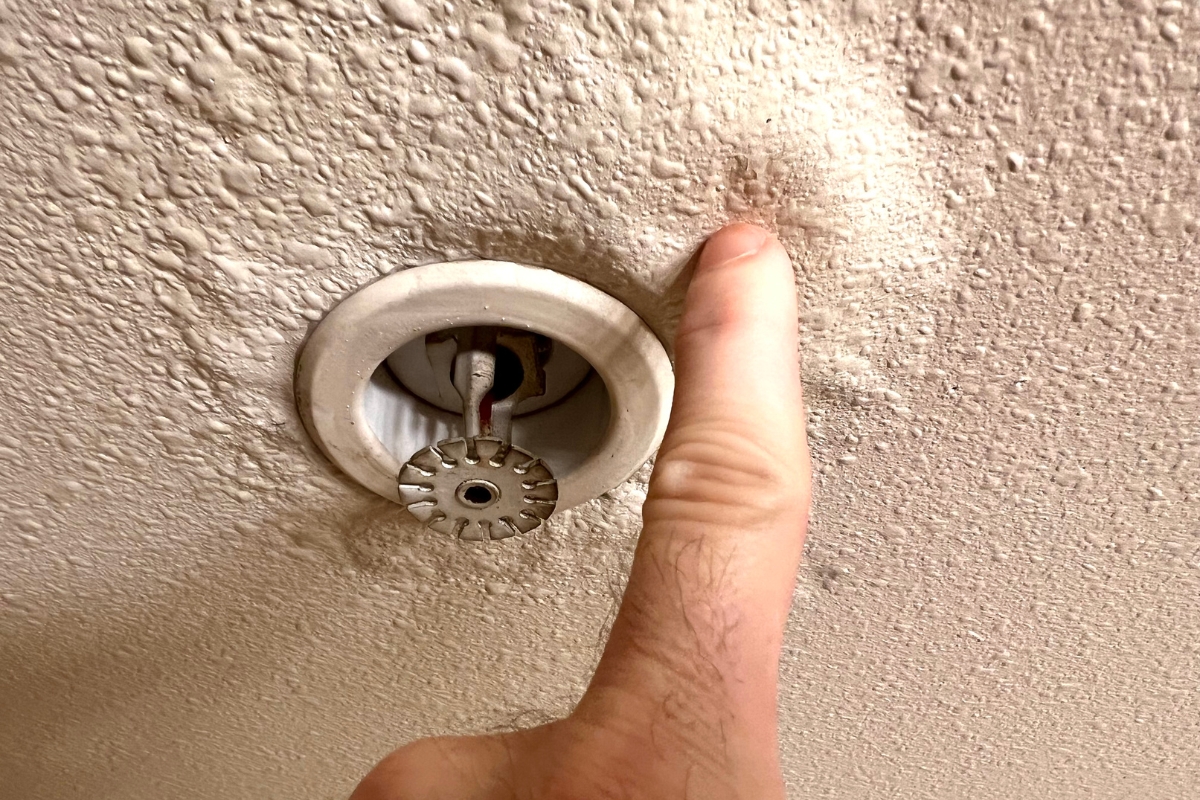 A hand points to a recessed fire sprinkler head mounted in a textured ceiling, highlighting a crucial component in preventing water damage.