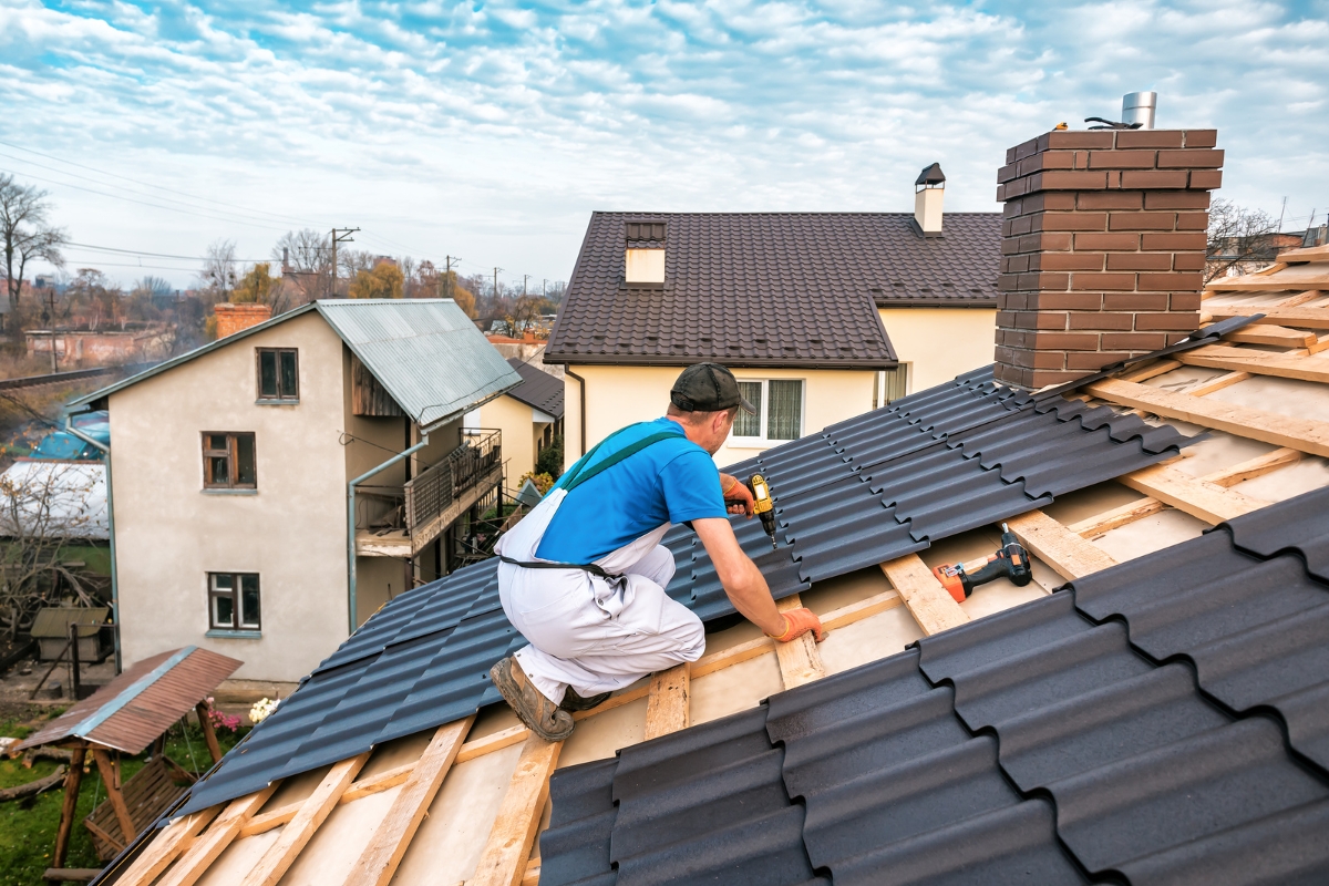 A worker is installing metal roofing panels on a residential roof, ensuring the structure is secure and preventing water damage, with other houses and a cloudy sky in the background.