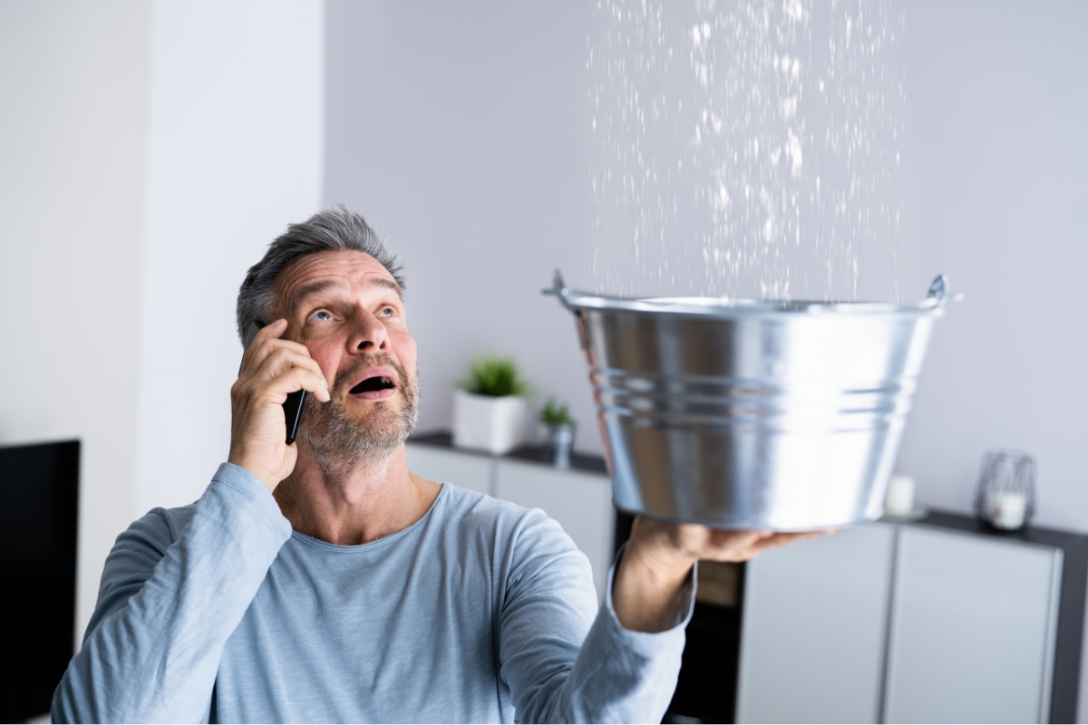 A man holds a metal bucket under a leak in the ceiling while talking on the phone, preventing water damage as streams of water cascade into the bucket.