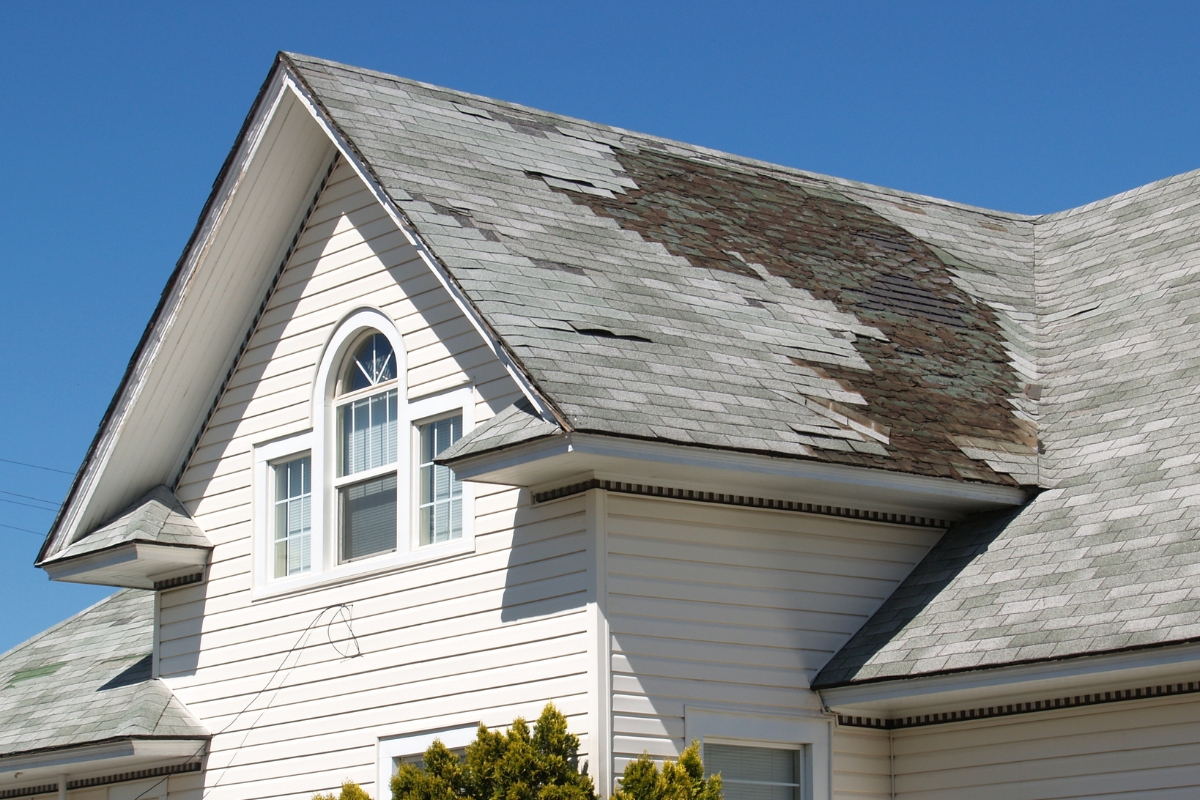 A house with white siding and a gable roof showing significant shingle damage on the right side under a clear blue sky, highlighting the importance of preventing water damage.