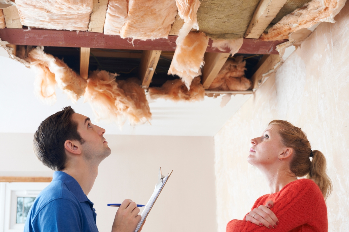 Two people inspecting a ceiling with insulation exposed, one holding a clipboard and pen, assessing the situation for water damage recovery.