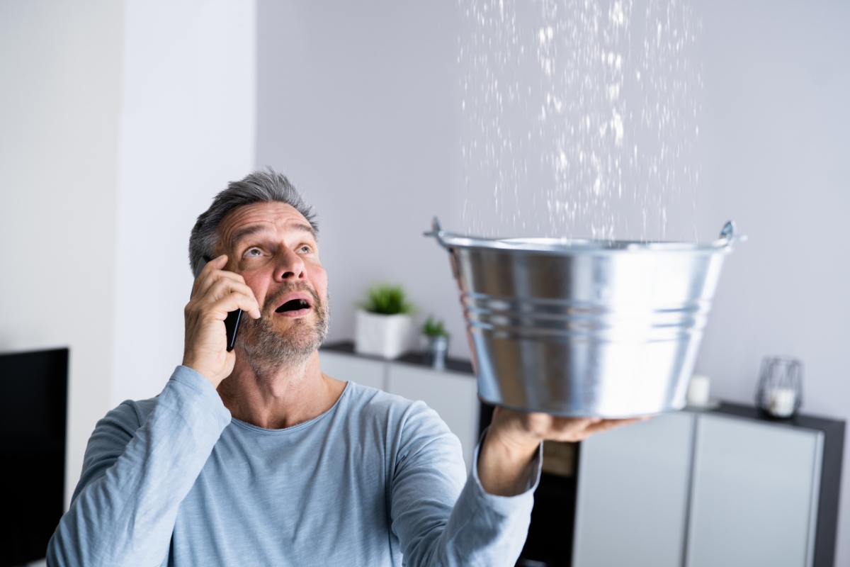 A man holds a bucket to catch water leaking from the ceiling while making a phone call, clearly worried about the situation. The scene is set in a minimally furnished room, hinting at the urgent need for water damage recovery.