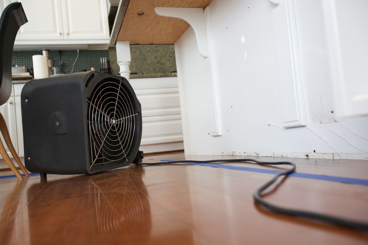 A black fan is positioned on the floor in a kitchen, blowing towards the base of a cabinet to aid in water damage recovery. The fan is connected to a power cord trailing along the wooden floor next to blue painter's tape.