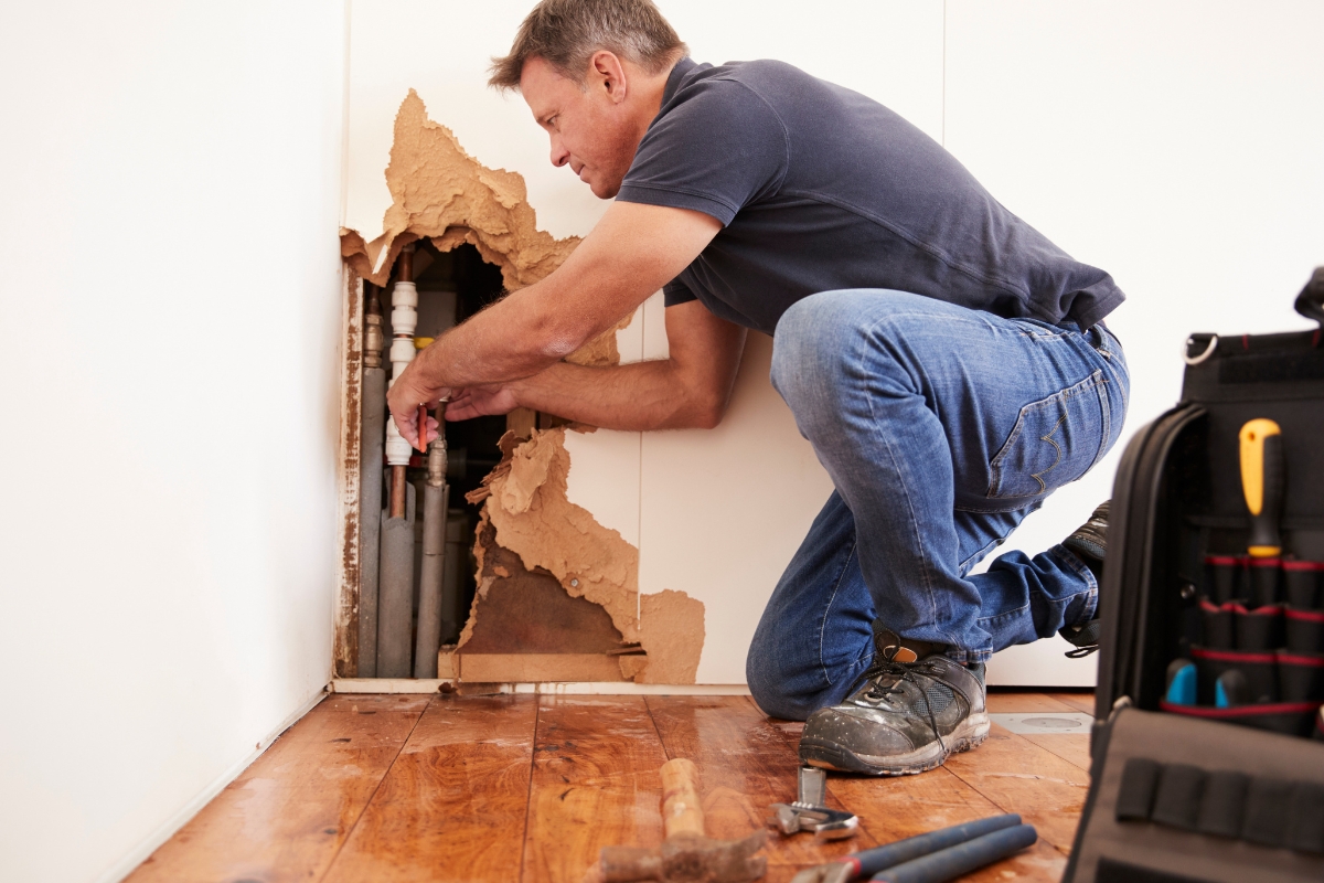 A man repairs plumbing behind a damaged wall, addressing water damage recovery, with tools scattered on the wooden floor and a tool bag nearby.