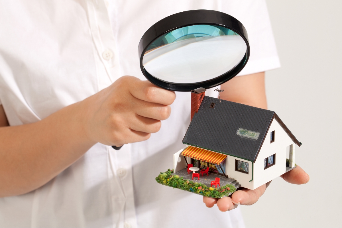 A person holds a magnifying glass above a small model house with an awning, red outdoor furniture, and signs of water damage recovery.