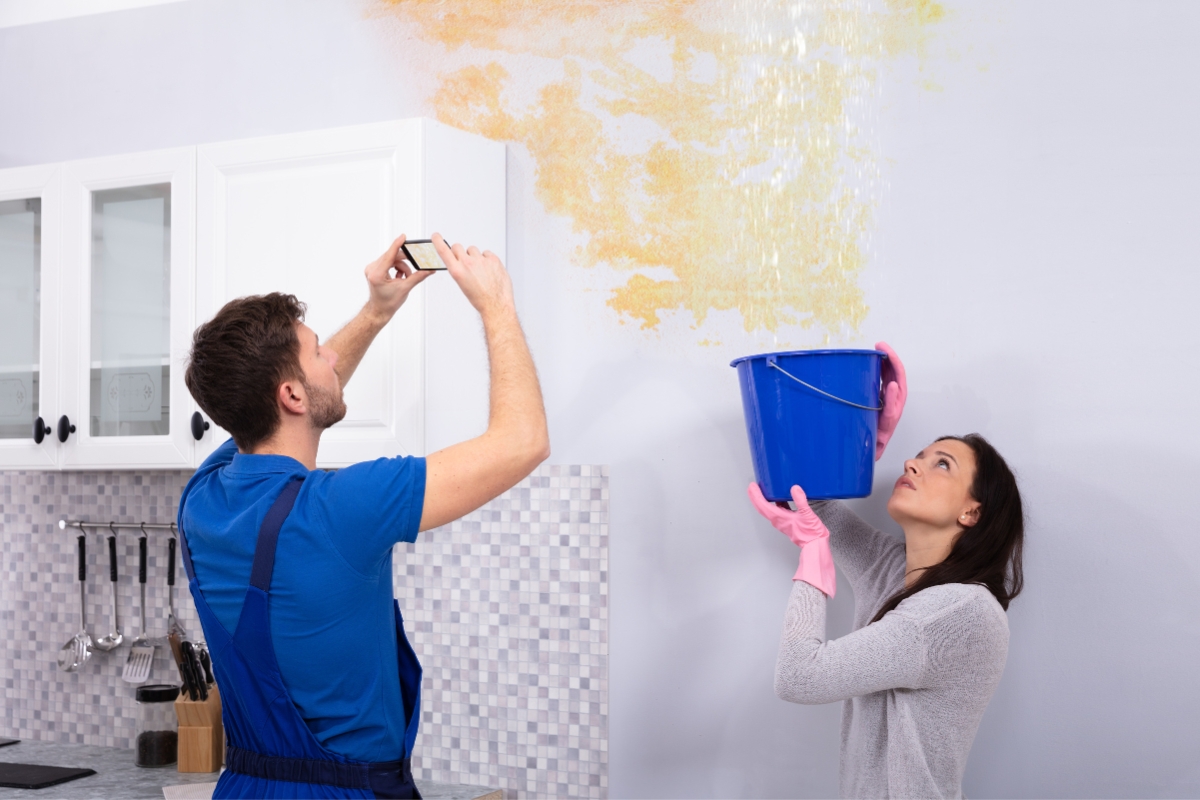 A man takes a photo of a ceiling leak in the kitchen while a woman holds a blue bucket to catch the water, both anticipating the next steps in their water damage recovery.