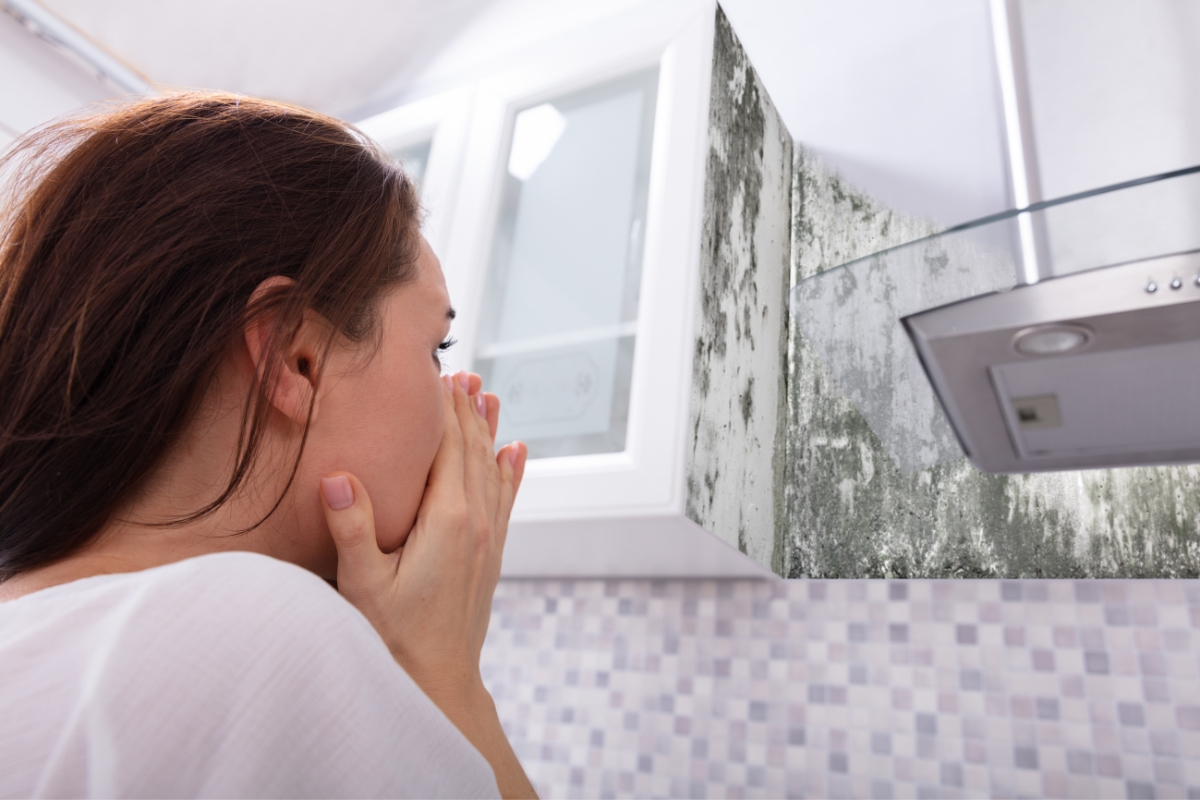 A person covers their mouth, alarmed at the kitchen wall near the oven hood covered in mold, aware of the health risks associated with mold.