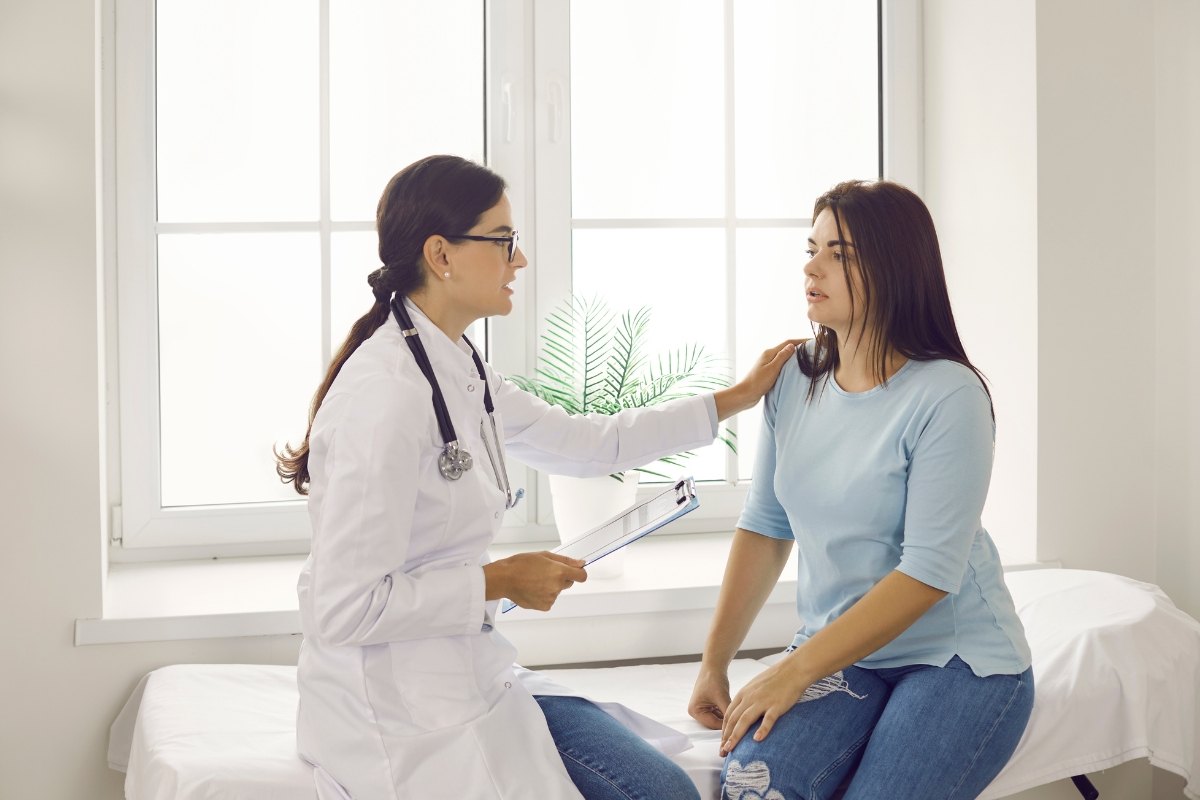 A doctor in a white coat with a stethoscope talks to a patient seated on an examination table. The doctor holds a clipboard and touches the patient's shoulder, discussing health risks associated with mold. They are in a brightly lit room with a window.