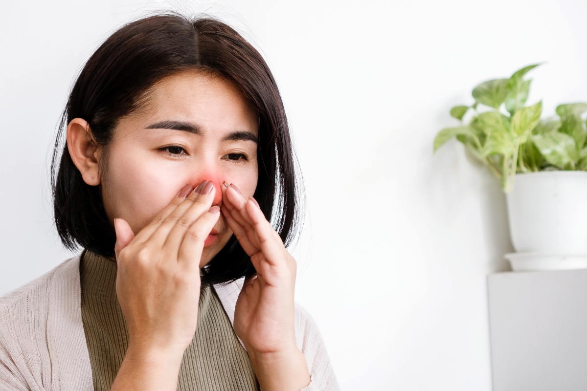 A woman with short black hair is holding her nose, appearing to be in discomfort, possibly due to health risks associated with mold, with a green potted plant in the background.