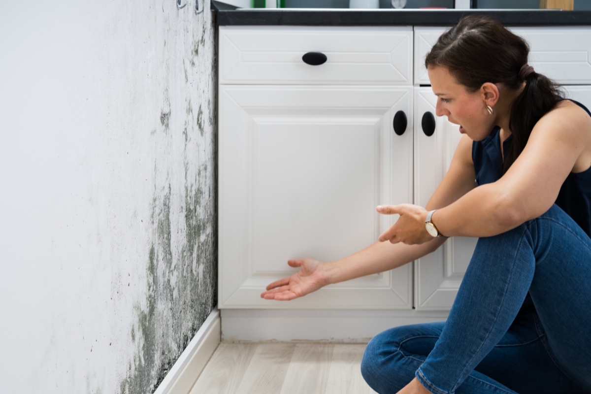 A woman in a kitchen points to a patch of black mold on a white wall near the base of the cabinets, highlighting the health risks associated with mold.