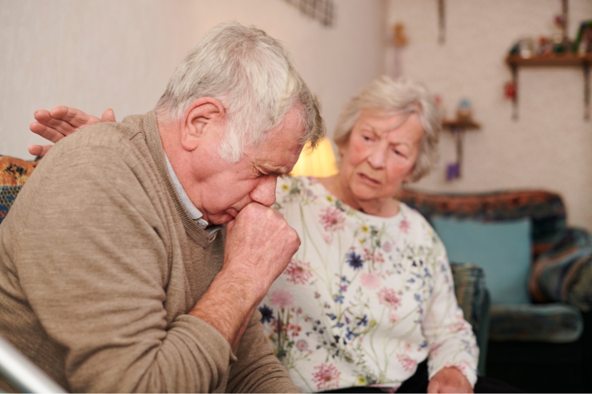 An elderly man sitting on a couch is coughing into his hand while an elderly woman beside him looks concerned and rests her arm on his back, worried about mold removal safety in their home.