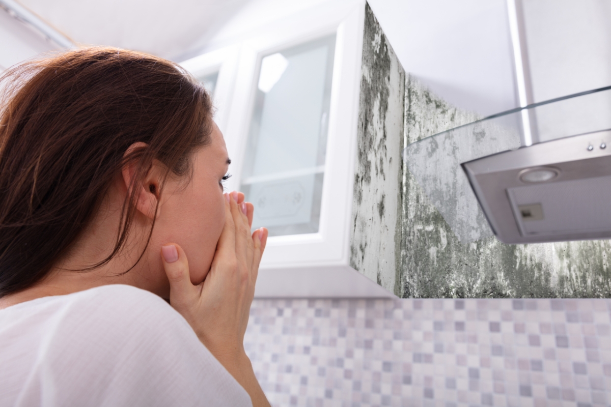 A woman covers her mouth in concern while looking at mold growing on a kitchen wall near a range hood, contemplating the importance of mold removal safety.