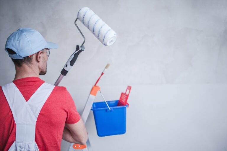 A man in a red shirt and white overalls, wearing a blue cap, holds a paint roller while standing beside a ladder with a blue bucket and paintbrushes, ready for surface preparation before painting.