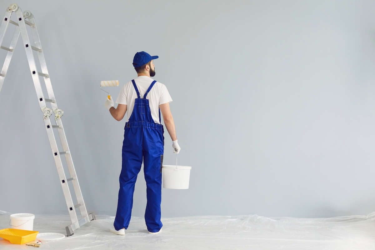 A person in blue overalls and a cap is painting a wall with a roller, having completed the necessary surface preparation before painting. They are holding a paint bucket and are next to a stepladder. The floor is covered with a protective sheet.