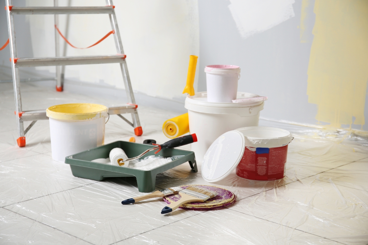 A room in the process of being painted showcases various painting supplies, including paint buckets, rollers, brushes, a ladder, and plastic sheeting on the floor—highlighting the importance of surface preparation before painting. The meticulous setup ensures a flawless finish.