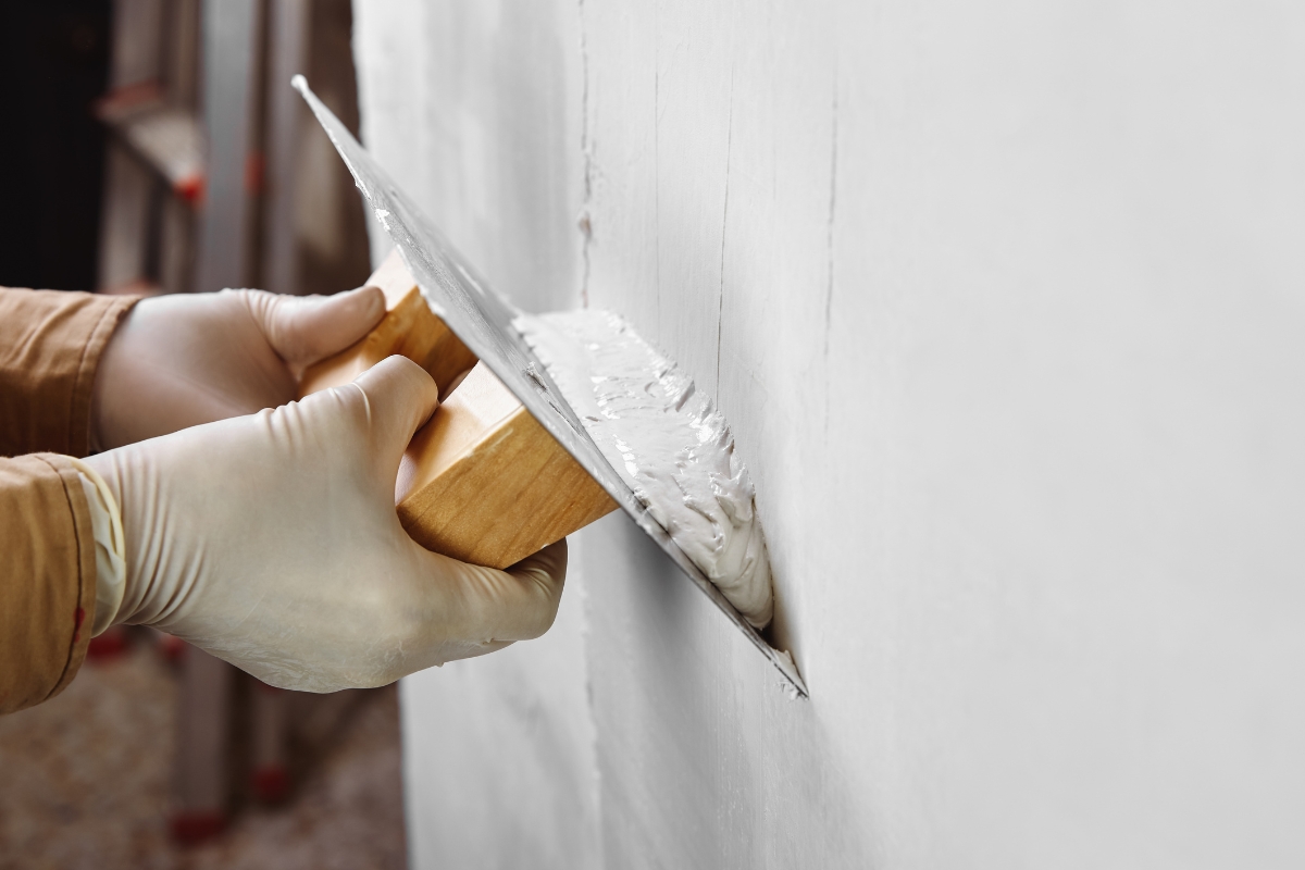 Close-up of a person with gloves applying plaster or putty to a wall using a spatula tool, focused on meticulous surface preparation before painting.