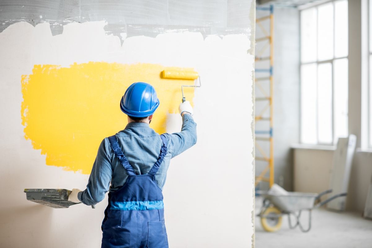 A worker wearing a blue hard hat and overalls is using a roller to paint a white wall yellow in a room under construction. Proper surface preparation before painting ensures the new color adheres smoothly. A scaffold and wheelbarrow are visible in the background.