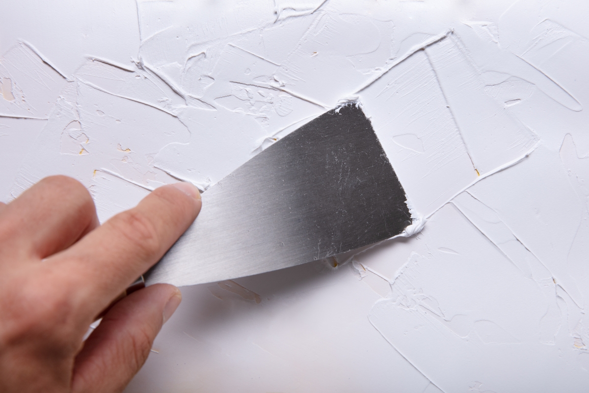 A hand smoothing white plaster on a wall with a metal putty knife is engaged in surface preparation before painting.