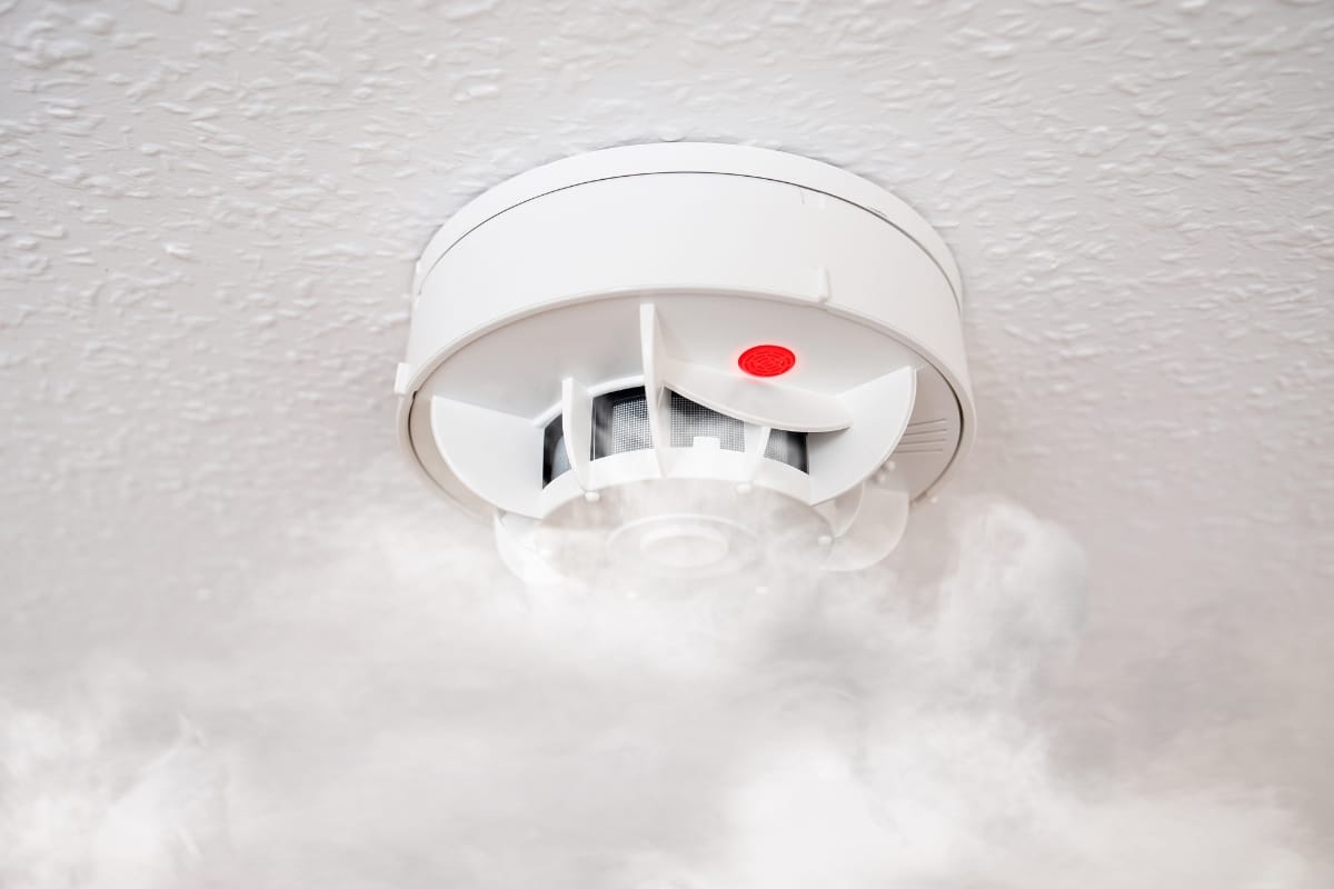 A smoke detector on the ceiling with a red light is surrounded by smoke, underscoring its vital role in fire prevention.