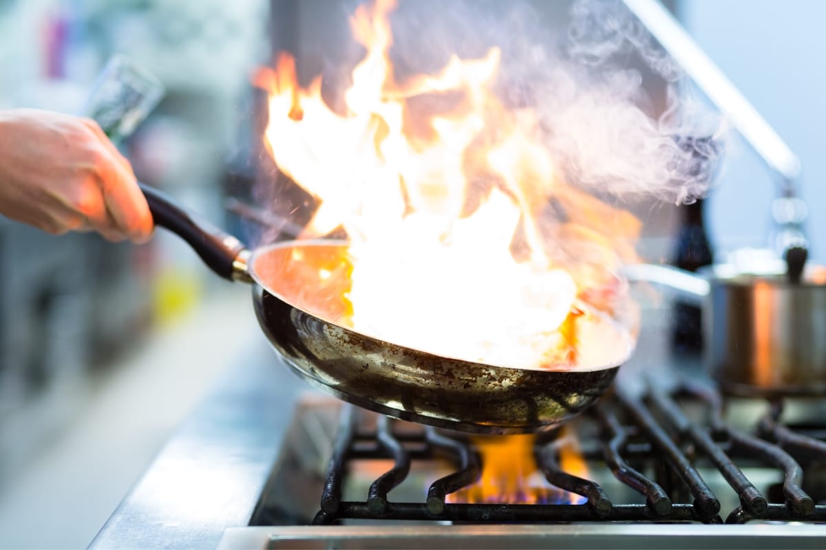 A person skillfully holds a frying pan with flames rising, over a gas stove in a kitchen setting, demonstrating keen awareness of fire prevention techniques to maintain safety.