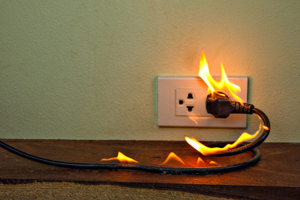 An electrical cord plugged into an outlet is on fire, with visible flames along the cord, highlighting the urgent need for effective fire prevention measures.