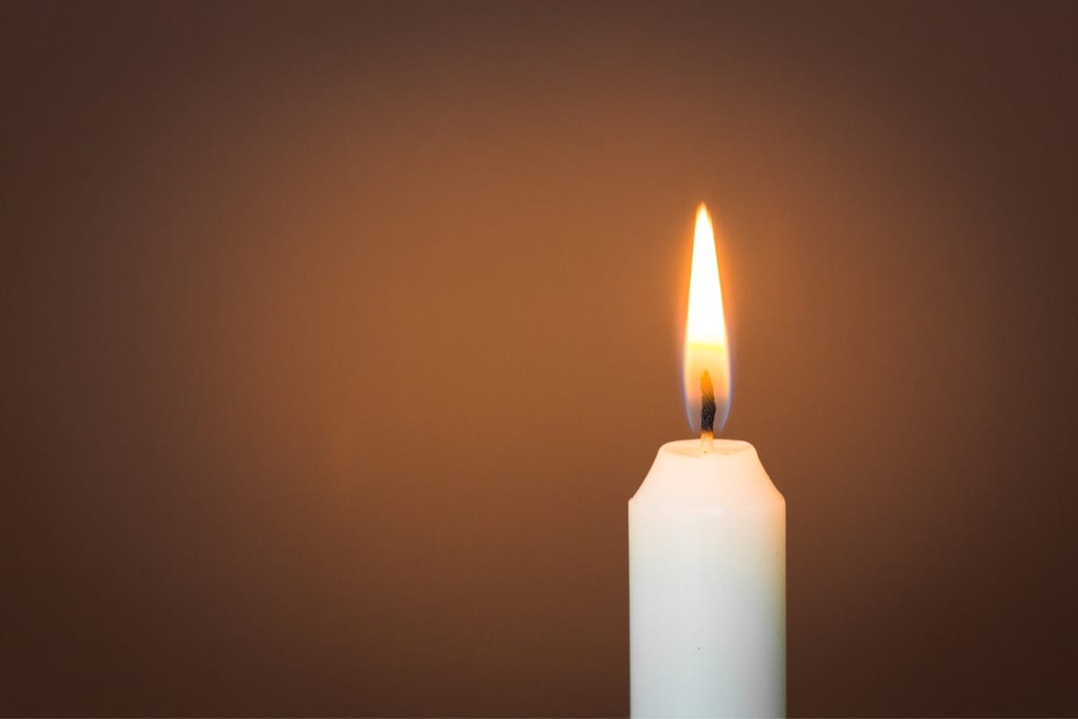 A single lit white candle flickers gently against a plain brown background, serving as a delicate reminder of the importance of fire prevention.