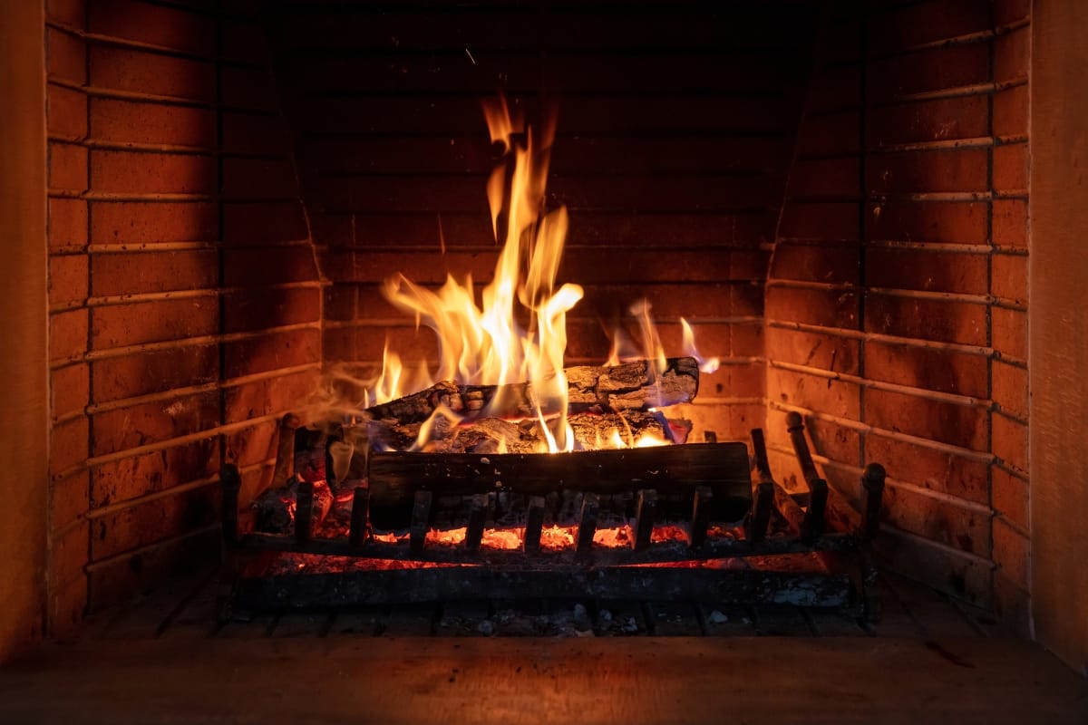 A lit fireplace with burning wood logs inside a brick hearth flickers warmly, while flames and embers create a cozy glow. Remember, practicing fire prevention ensures that this comforting scene remains safe and enjoyable.