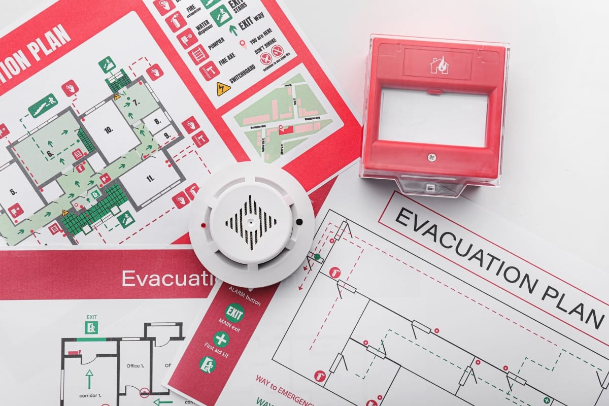 Evacuation plans, a smoke detector, and a fire alarm box are displayed on a white surface, showcasing essential elements of fire prevention.