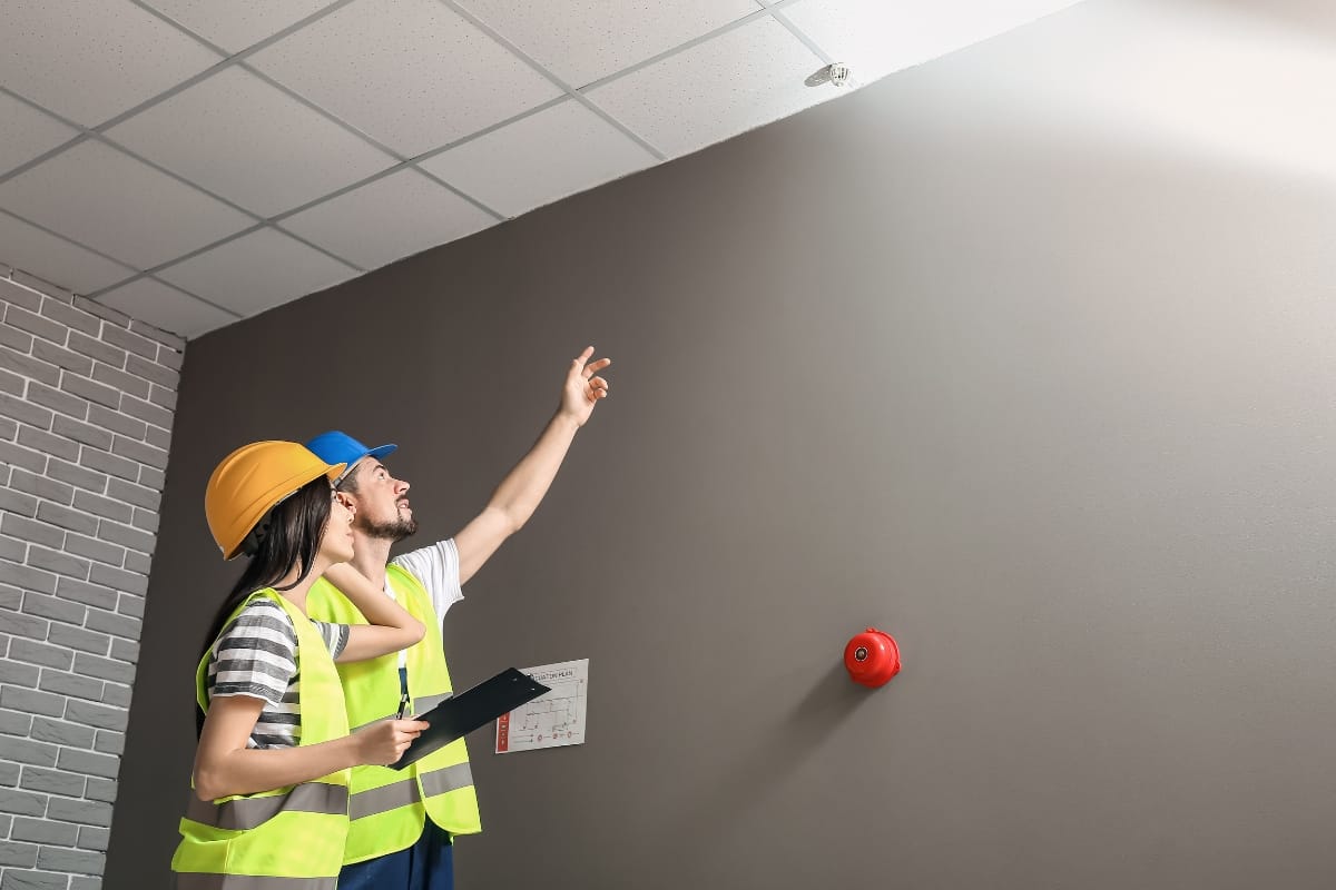 Two workers in safety vests and helmets inspect a ceiling with a keen focus on fire prevention. One points upwards while the other holds a clipboard, noting observations. A fire alarm is mounted on the wall, underscoring their commitment to maintaining safety and vigilance.